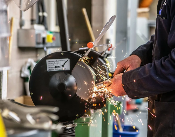 Close-up of a grinding wheel in action, with sparks flying as a lecturer in a navy-blue coat sharpens a metal component. Close-up of a grinding wheel in action, with sparks flying as a lecturer in a navy-blue coat sharpens a metal component.
